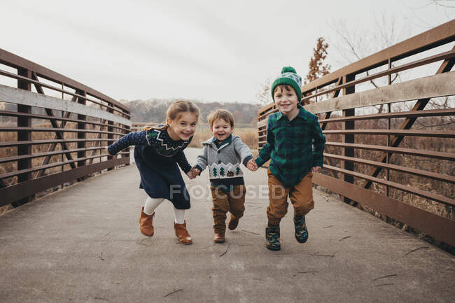 Трое братьев и сестер держатся за руки, бегут по мосту к камере — стоковое фото