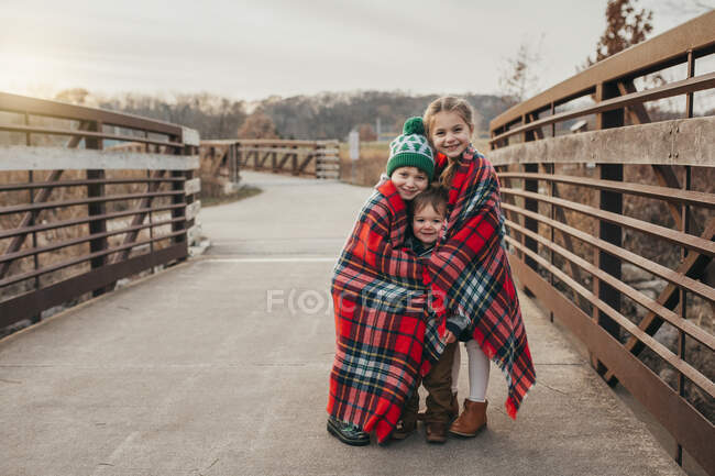 Siblings wrapped in Christmas plaid blanket on bridge at sunset — Stock Photo