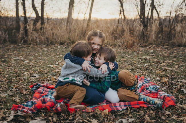Tres hermanos sentados en manta a cuadros roja abrazándose al atardecer - foto de stock