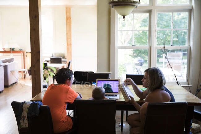 Мама и сын Тоддлера смотрят ноутбук сидеть с кузеном-подростком за столом — стоковое фото