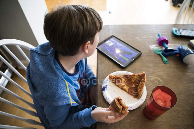Вид сверху на мальчика-подростка с гриппом, поедающего пиццу, смотрящего Ipad за столом — стоковое фото