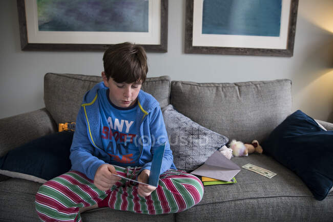 Мальчик-подросток в полосатой пижаме читает открытку на день рождения, сидя на диване — стоковое фото