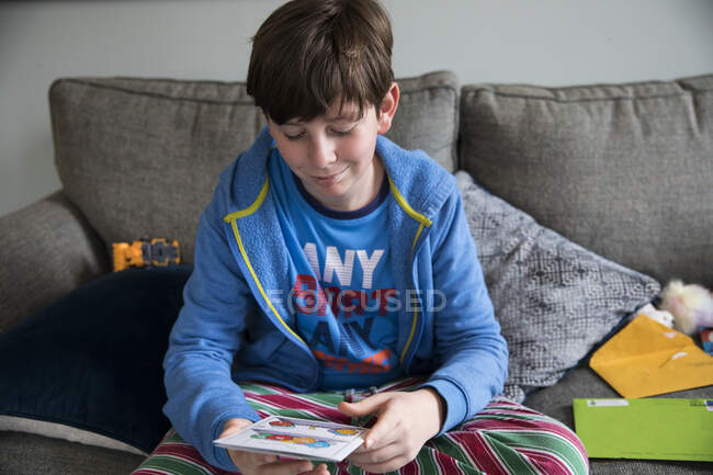 Lächelnder Teen Boy liest Geburtstagskarte, während er auf Couch in Pjs sitzt — Stockfoto