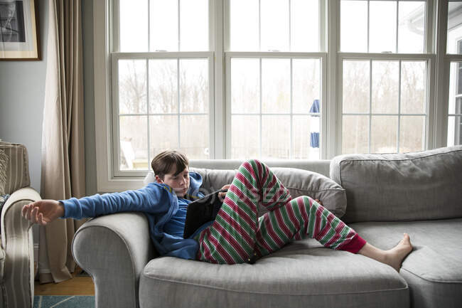 Adolescente com gripe, em pijamas listrados, senta-se no sofá assistindo Ipad — Fotografia de Stock