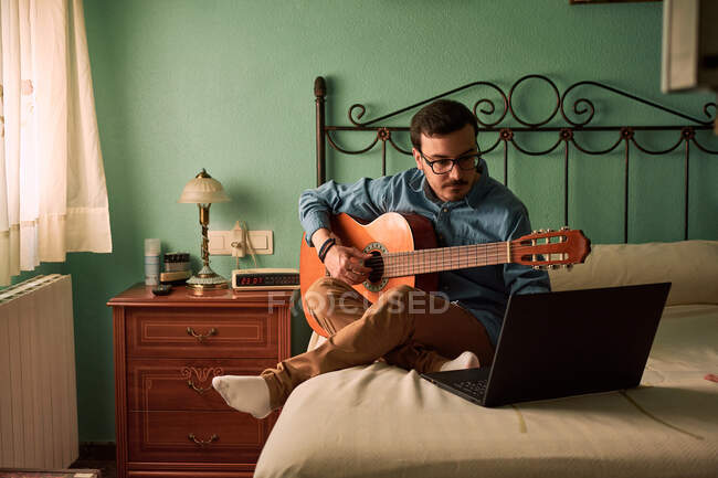 Молодий чоловік практикує з гітарою, дивлячись на свій ноутбук на — стокове фото