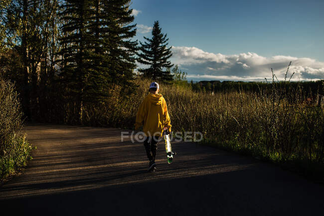 Adolescente chico caminando al atardecer con monopatín - foto de stock