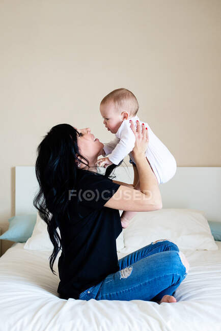 Charmante femme brune jouant avec son bébé tout en étant assise sur le lit — Photo de stock