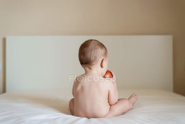 Bebé desnudo sentado en la cama sobre su espalda sosteniendo una manzana - foto de stock
