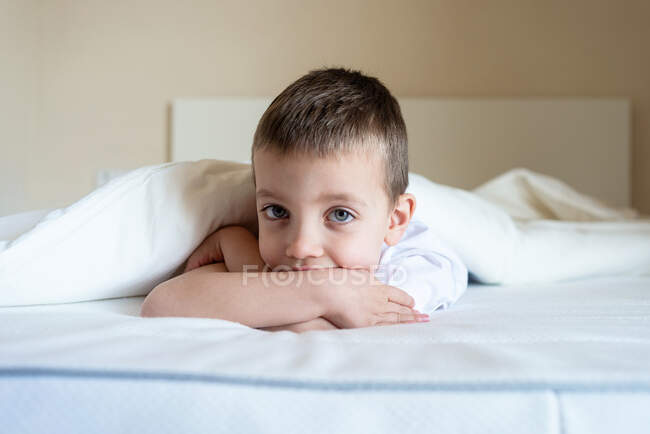 Liebenswertes Kind, das unter einer Decke auf dem Bett liegt, während es in die Kamera schaut — Stockfoto