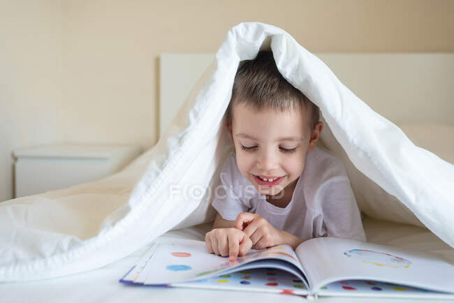 Adorable niño sonriente acostado en la cama debajo de la manta, leyendo un libro - foto de stock