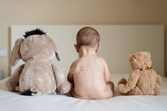 Rear view of a naked baby sitting up in bed with two teddy bears — Stock Photo