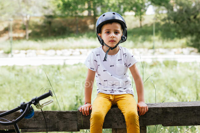 Ragazzo di 4 anni seduto su una recinzione di legno con la sua custodia per bici su uno sfondo verde di fronte alla fotocamera — Foto stock