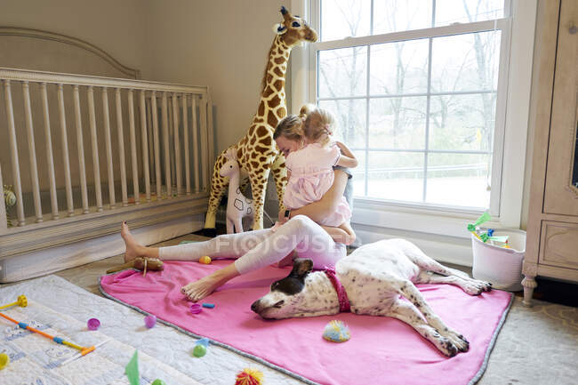 A mom and daughter hug in child's bedroom, dog lays beside them — Stock Photo