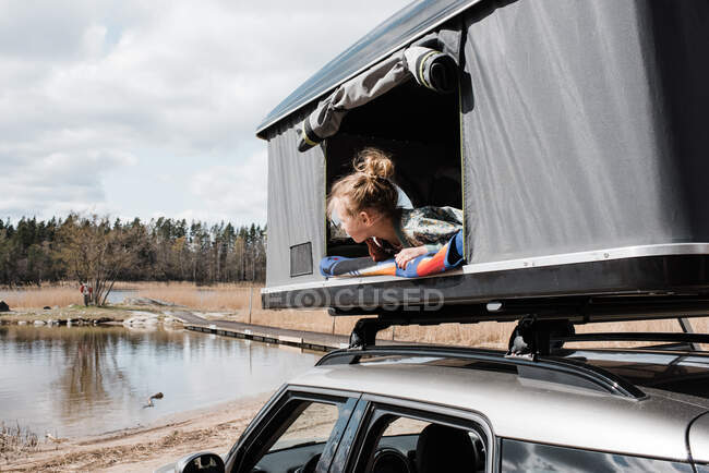 Girl looking out of roof top tent at a view of a lake while camping — стоковое фото