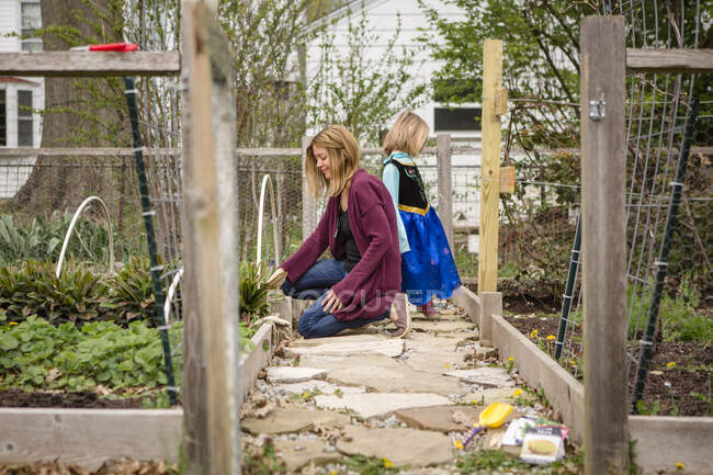 Uma mãe trabalha em seu jardim enquanto uma criança de traje aguarda — Fotografia de Stock