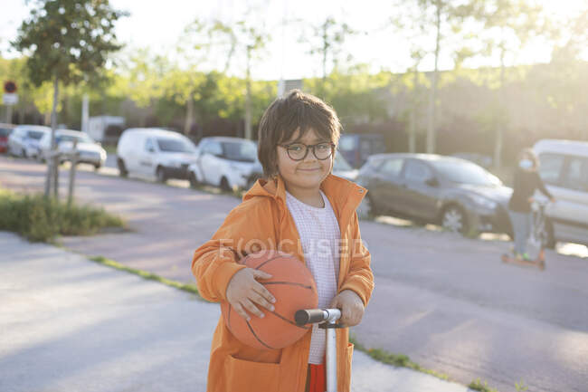 Jeune garçon avec une balle de basket chevauchant un scooter sur un trottoir — Photo de stock