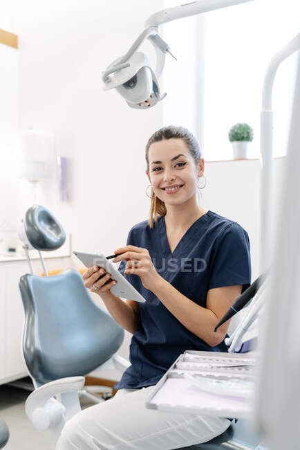 Молода медсестра в синій куртці посміхається в стоматологічному кабінеті, перш ніж приймати пацієнта і обробляє планшет . — стокове фото