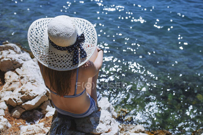 Vista trasera de una mujer con sombrero sentada junto al mar - foto de stock
