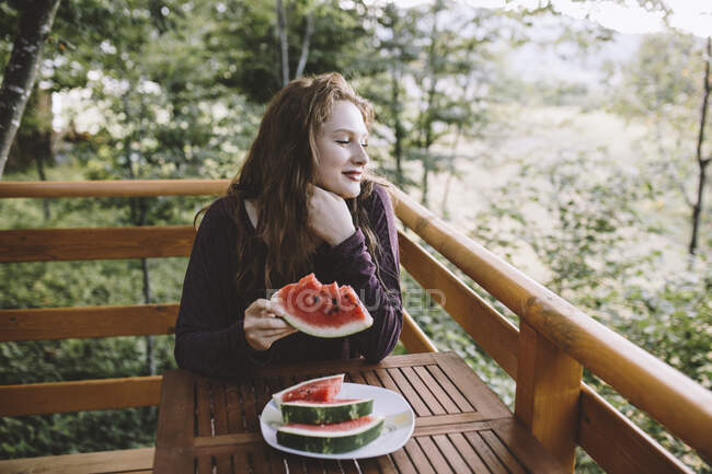 Молодая рыжая женщина ест арбуз на балконе деревянного дома — стоковое фото