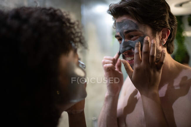 Mulher afro-americana desfocada ajudando namorado alegre a manchar argila no rosto durante o procedimento de spa em casa — Fotografia de Stock