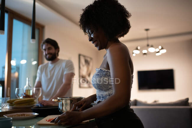 Vista lateral da mulher afro-americana feliz sorrindo e cortando ingredientes para o jantar antes da data romântica com o namorado em casa — Fotografia de Stock