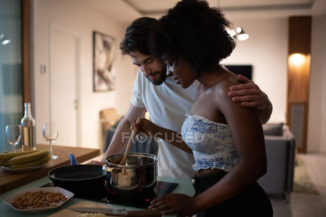 Bearded young man embracing African American woman and looking into saucepan while cooking romantic dinner in cozy kitchen together — Stock Photo