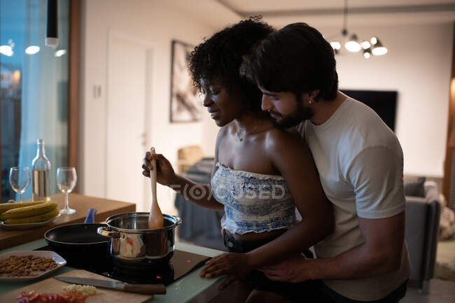 Barbudo cara abraçando afro-americano mulher por trás enquanto cozinhar jantar romântico juntos à noite em aconchegante apartamento — Fotografia de Stock