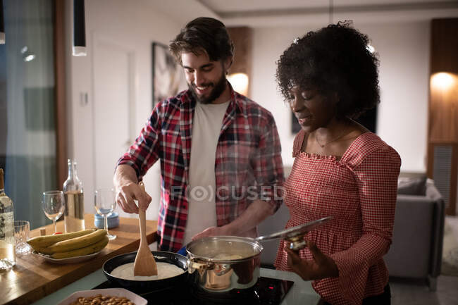 Feliz hombre y mujer multirracial en ropa casual sonriendo y cocinando deliciosos platos juntos antes de la cena romántica en casa - foto de stock
