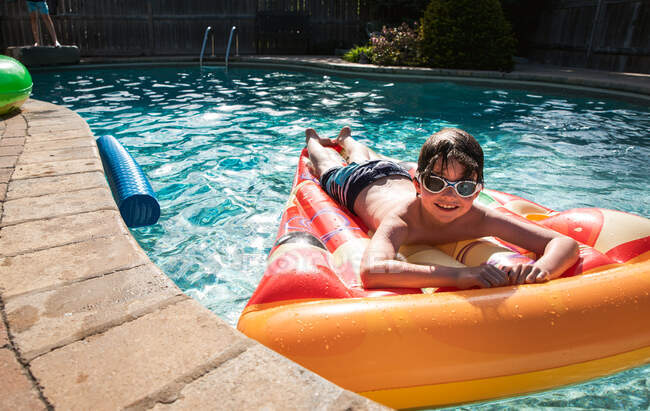 Niño joven con gafas en un flotador de pizza en una piscina patio trasero. - foto de stock