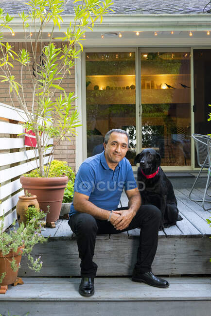 Sonriente macho hispano sentado en su cubierta con perro negro - foto de stock