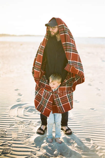 Отец греет своего сына одеялом на ветреном пляже — стоковое фото