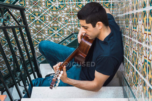 Young man playing guitar sitting on a staircase with beautiful tiles. — Stock Photo