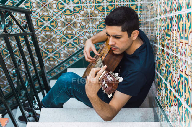 Focus man playing guitar sitting on a staircase with beautiful tiles. — Stock Photo
