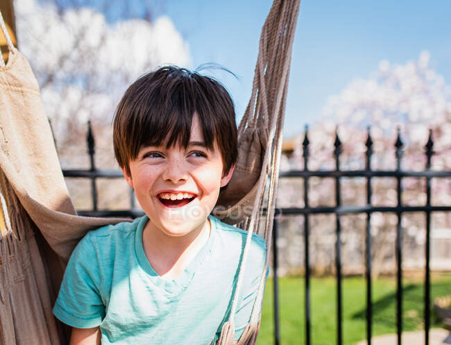 Niño feliz sentado en una hamaca en un patio trasero en un día de verano. - foto de stock