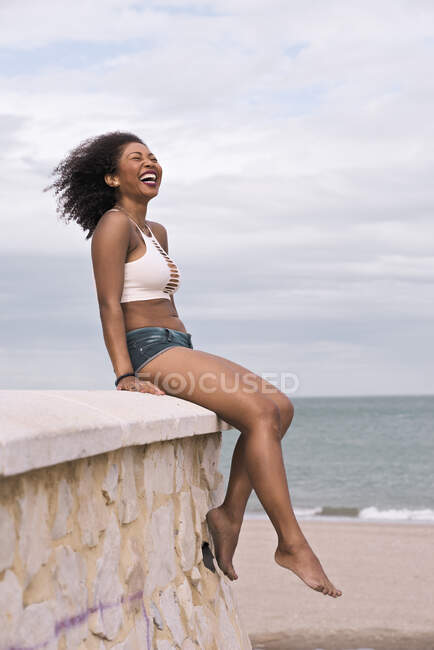 Young black woman laughs sitting on top of a stone wall by the sea — Stock Photo
