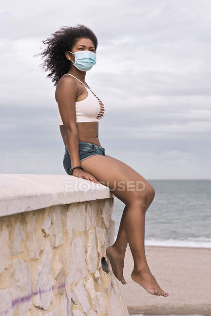 Mujer negra joven contempla el mar sentado en la parte superior de un muro de piedra, protegido por una máscara. Covid-19 - foto de stock