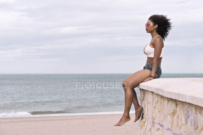 Молодая черная женщина созерцает море, сидящее на каменной стене — стоковое фото