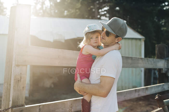 Um pai segura sua filha em uma fazenda em uma noite ensolarada. — Fotografia de Stock
