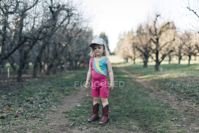 Une jeune fille se tient dans un verger portant un justaucorps et des bottes de cowgirl. — Photo de stock