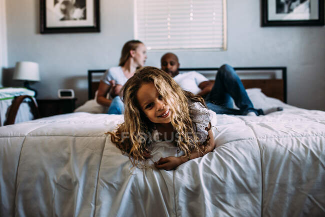 Giovane ragazza che gioca alla fine del letto con i genitori in background — Foto stock