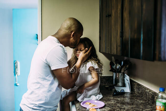 Папа целует дочь, пока она сидит на кухонном столе — стоковое фото