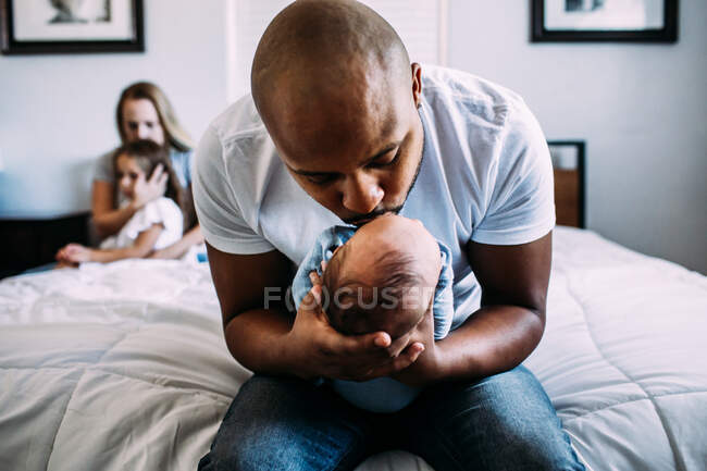 Dad kissing newborn on bed with mom and daughter in back ground — Stock Photo