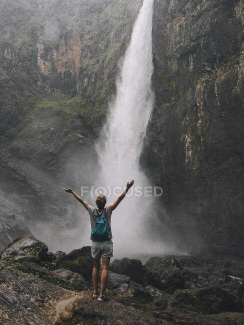 Visão traseira de uma jovem mulher abrindo os braços enquanto olha para a cachoeira, Queensland, Austrália. — Fotografia de Stock