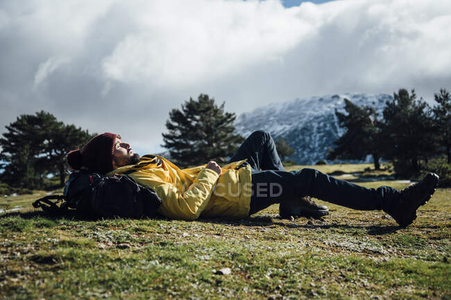 Молодой человек в желтой куртке и рюкзаке лежит на горе. — стоковое фото