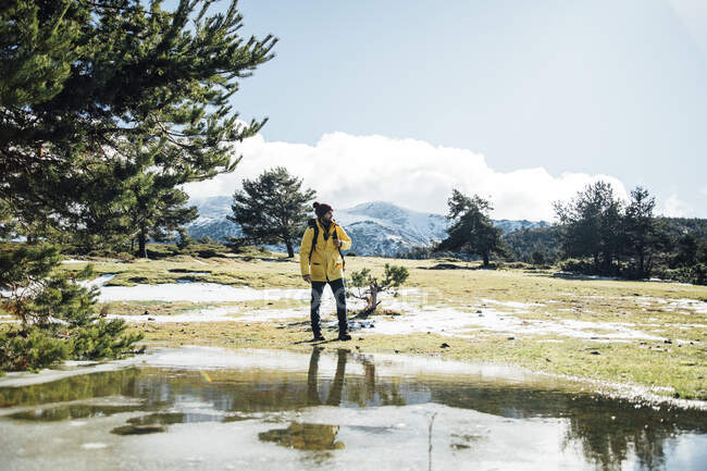 Молодой человек в желтой куртке и рюкзаке рядом с прудом в горах. — стоковое фото