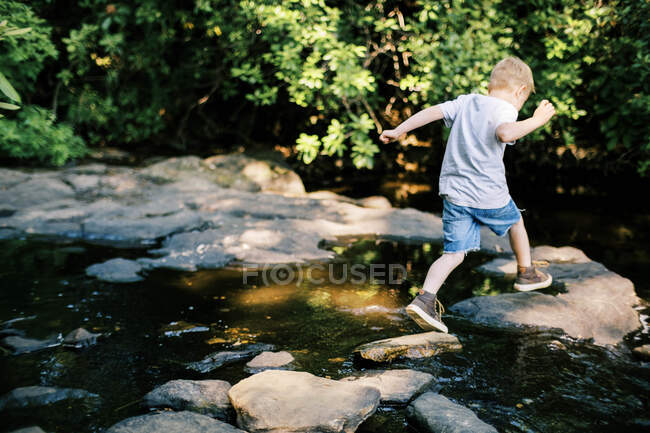 Un brave garçon de 5 ans sautant par-dessus des rochers dans une rivière — Photo de stock
