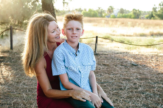 Ein süßer Junge mit roten Haaren sitzt draußen auf dem Schoß seiner Mutter — Stockfoto