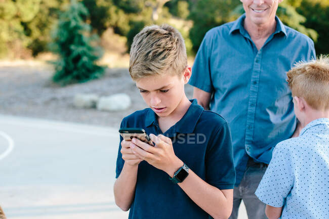 Мальчик-подросток смотрит на свой смартфон в то время как снаружи с семьей — стоковое фото