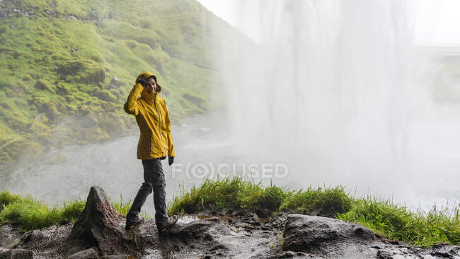 Joyeux randonneur féminin dans un imperméable lumineux derrière une cascade puissante — Photo de stock