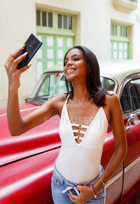 Joven cubano tomando una selfie frente a un auto viejo en La Habana, Cuba - foto de stock